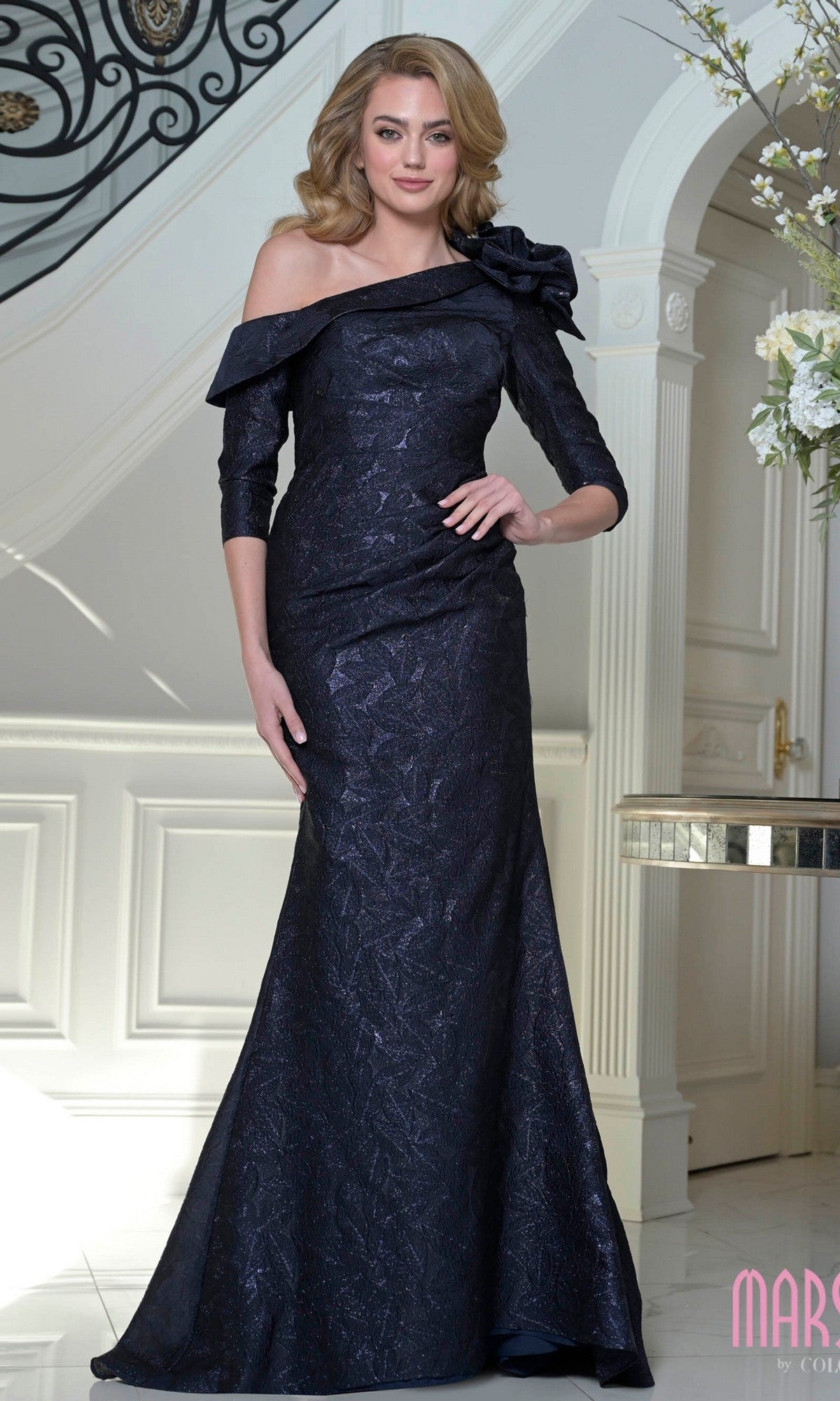 A woman in the Marsoni MV1399 long formal dress in navy, featuring jacquard fabric and a floral shoulder accent, stands elegantly by a staircase in an opulent interior.